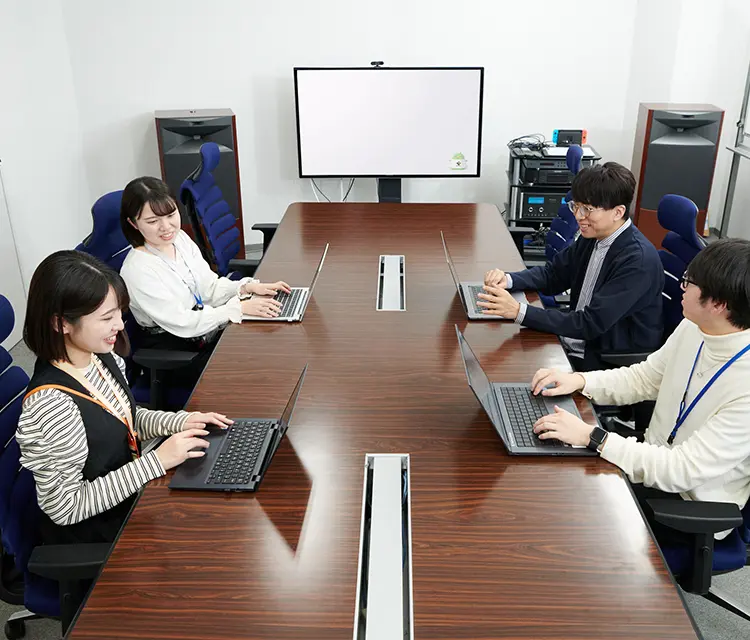 写真：会議テーブルで各自ノートPCを扱いながら打ち合わせをしている男性社員2名と女性社員2名